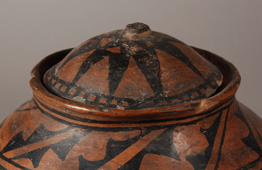 Tesuque Pueblo Black-on-red Jar with Lid Tesuque Black-on-red pottery is fashioned in the same manner as polychrome wares. Clay, temper, paste, and construction do not differ. The difference arises when a red slip is wiped on the vessel in lieu of a cream slip. The underside of the vessel is concave, the vessel body is rounded in the best tradition of Tewa vessels, and the design is sparse, all of which point to a date of origin of circa 1880. The light tan paste at the lower third of the vessel exhibits the bumpiness that is a trademark of Tesuque. A red band at the upper end of the tan paste divides the design area from the underbody. Double framing lines above this red band, at the shoulder, and just below the rim serve to isolate the two design areas. The mirrored C-like design with eye-like elements and typical chevron elements cleverly conceals two Avanyu (Water Serpents) with their heads pointing down. The inner rim of the jar is designed to support a lid. It was not unusual at Tesuque for the potters to make lidded jars. Most lids have been lost or broken. Fortunately the lid for this jar is still here, missing only the knob.