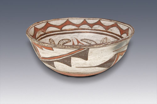 Occasionally we acquire a piece of pottery that is a joy to hold and a visual delight. Such is this marvelous Zuni Pueblo serving bowl. Not only was it constructed by a master potter and painted by a talented artist, it was perhaps lovingly used in the pueblo for decades before someone talked the owner into parting with it. It dates to the last half of the nineteenth century. The paste materials are typically Zuni; white and chunky in texture with the centuries old temper material of ground pottery shards. The use of old shards for temper evidences the regard of the pueblo peoples for the old works made by their predecessors. To add these remnants to the new work is a sign of respect.   The rim of the bowl flares outward slightly, imparting a graceful shape to what could be an otherwise uninteresting one. The bowl is slipped in traditional fashion with cream-colored clay, over which is painted the design in mineral and vegetal paints. The underbody is slipped in red, a tradition generally abandoned in the mid-1860s and completely abandoned by 1880. The exterior is decorated with the traditional prayer stick design used on every Zuni bowl made in the nineteenth century. It is thought that this was a woman's way to place her prayers since women do not make prayer sticks.   The interior decoration begins with a brown rim, below which is a wave-like element encircling the bowl that is painted deep red and outlined in brown. Below this are two wide parallel framing lines with a very distinctive ceremonial break. The main body of the interior is elaborately designed with polka-dotted floral elements and red Zuni volute elements without the addition of brown outlining. The base of the bowl features a red square, outlined in brown, which is again outlined with a wavy brown frame.   This is one of the most extraordinary bowls we have had the pleasure to handle. A two-square-inch piece of clay blew out during firing, but the potter placed it back in and “cemented” it in place so that she could still use the bowl for home use. The piece has been professionally repaired at that point of damage. Provenance: Cody Wyoming Collection. This Zuni Serving Bowl was part of the exhibit CODY, WYOMING POTTERY COLLECTION June 10, 2006 until September 10, 2006 and BOWLS: A Pueblo Necessity December 15, 2006 until December 31, 2006 presented at Adobe Gallery Santa Fe.