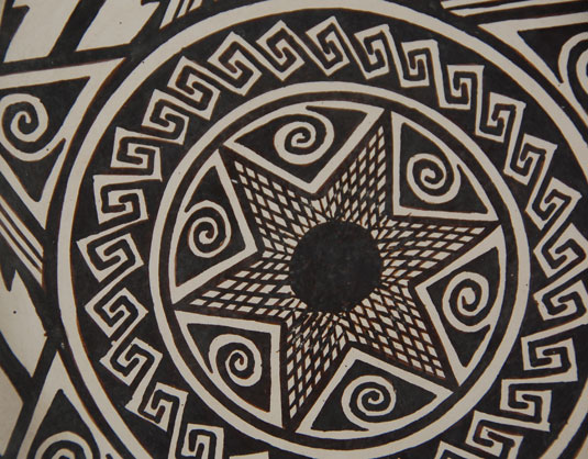 .  The design is a mixture of shapes—a star in the center surrounded by circles, one of which contains Greek Key-style designs.  Beyond the circles are triangles with spirals, triangles with cross hatching, and black parallelograms.  The exterior rim has a band of cloud symbols interspersed with black dots.  